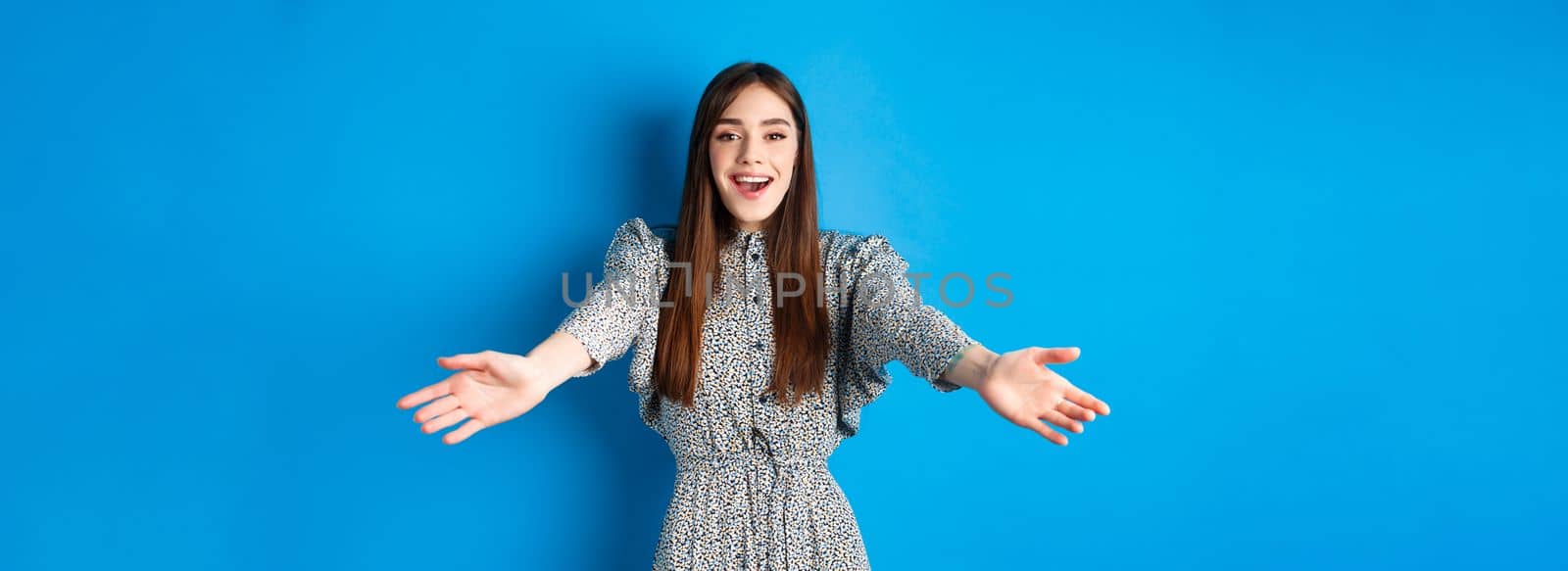 Beautiful happy woman in dress stretch out hands in warm welcome, smiling and greeting you, inviting guests, standing on blue background.