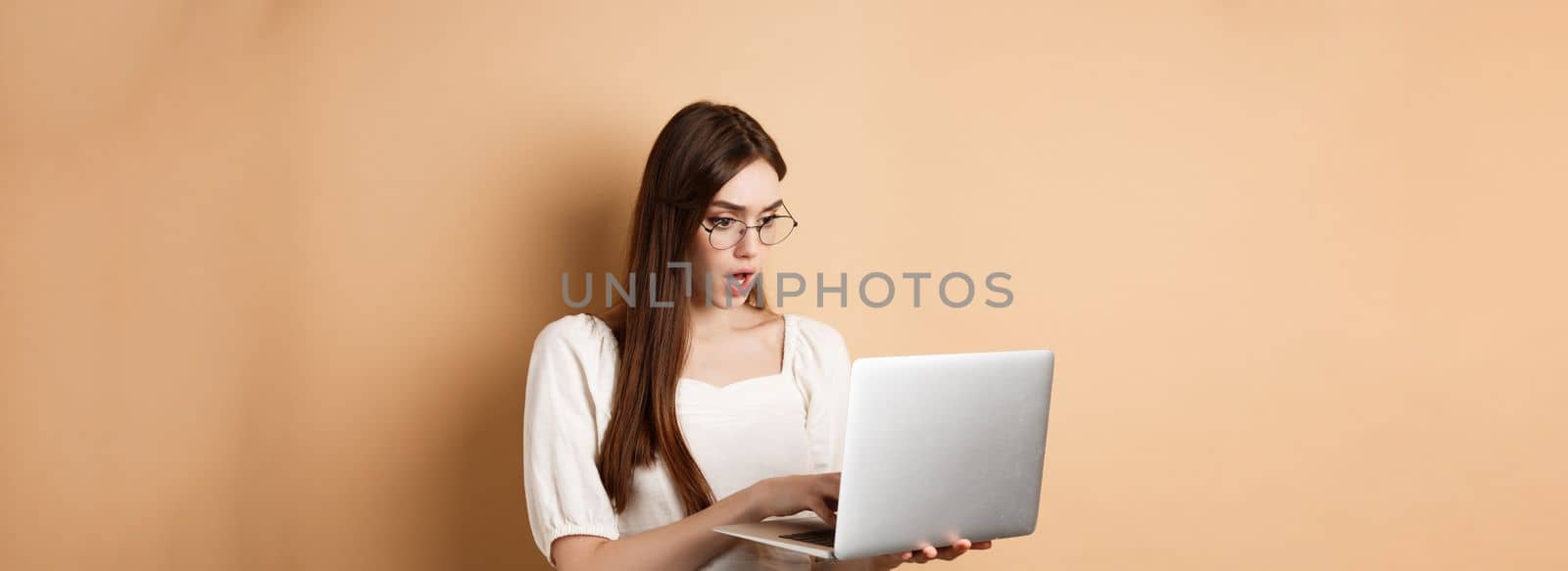 Worried girl look at laptop screen, typing on computer with focused face. Woman freelancer working in internet, standing on beige background.