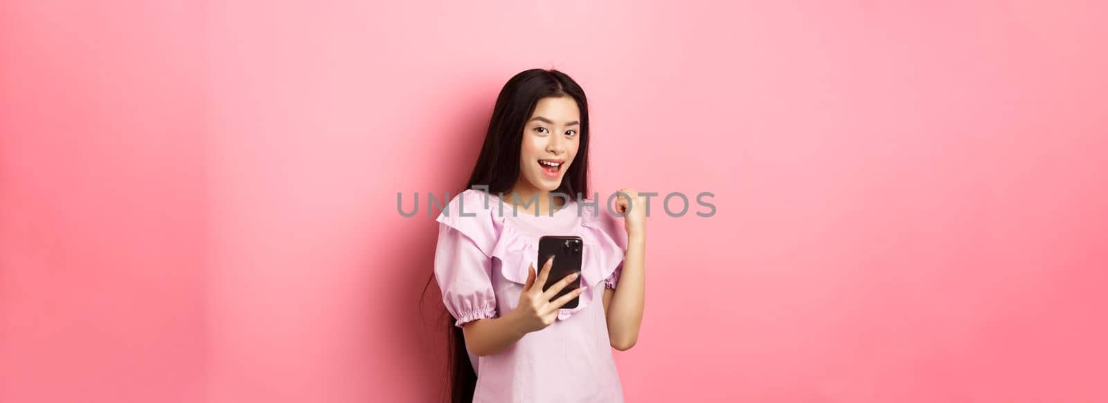 Online shopping. Satisfied asian girl winning on mobile phone, say yes and make fist pump, holding smartphone, standing against pink background.