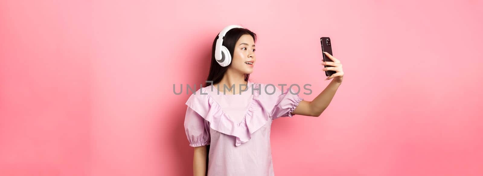 Beautiful asian girl blogger taking selfie in wireless headphones, make photo for social media on smartphone, standing in dress against pink background by Benzoix