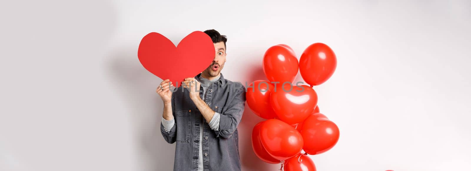 Cute young man hiding face behind big Valentine heart card, secret admirer looking at camera with amazement, saying wow, standing near romantic balloons on white background by Benzoix