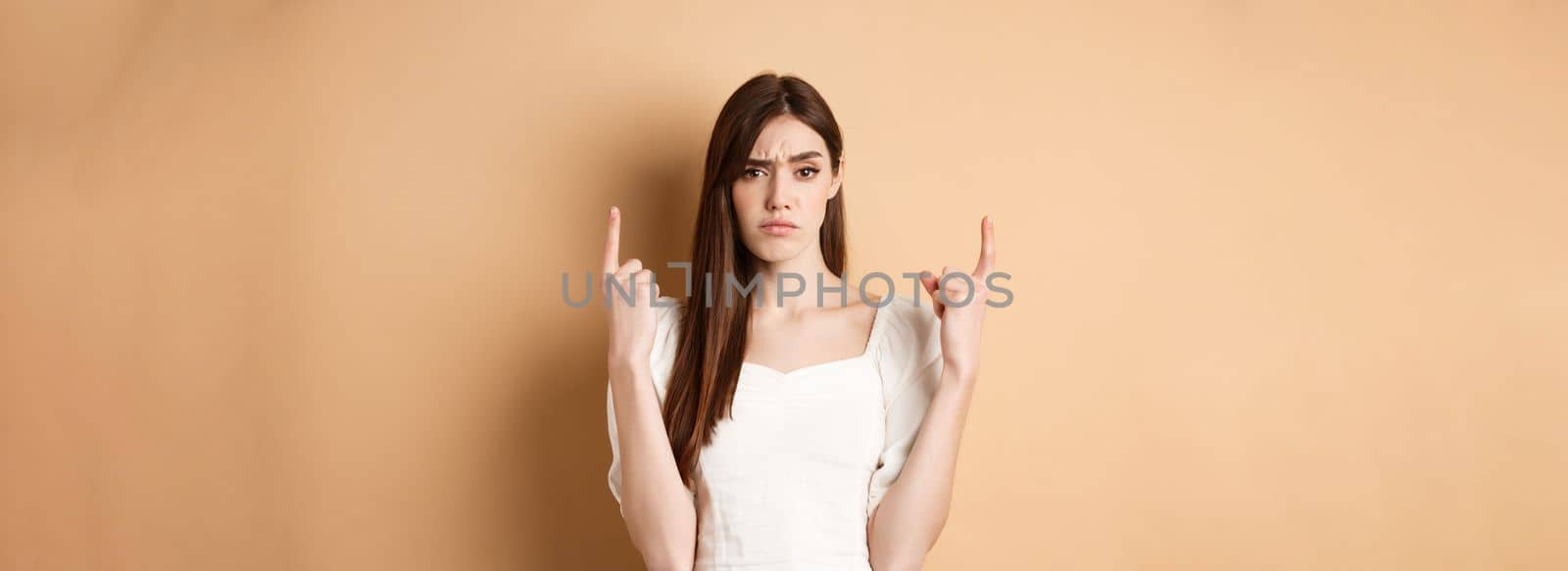 Skeptical woman frowning and pointing fingers up, having doubts about product, standing on beige background by Benzoix