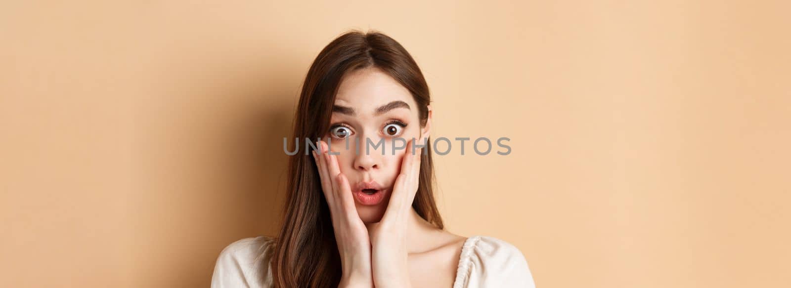 Face of surprised girl saying wow, touching cheeks and staring excited at camera, standing on beige background by Benzoix