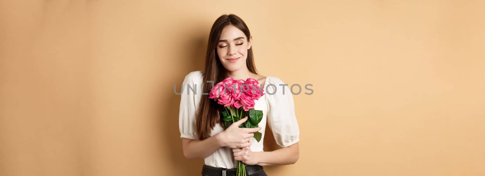 Valentines day. Tender and romantic caucasian woman close eyes dreamy, hugging bouquet of roses from secret admirer or lover, standing on beige background by Benzoix