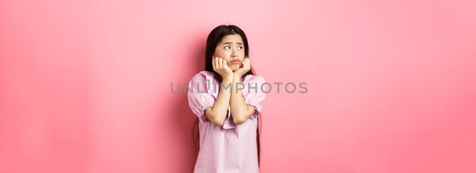 Sad and lonely asian girl look left and frowning, standing distressed against pink background.