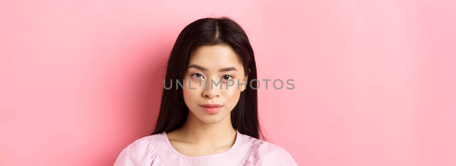 Close-up of beautiful young asian woman in dress, looking confident at camera, standing on pink background.