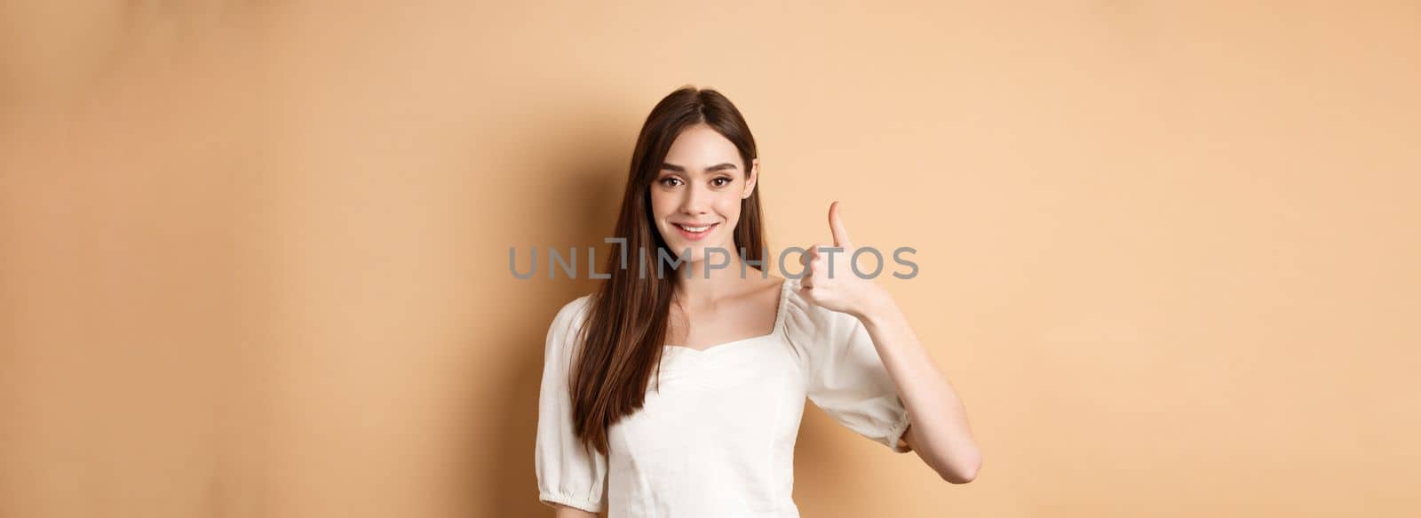 Good job. Smiling candid woman in blouse showing thumb up, praise nice choice, recommending product, standing satisfied on beige background by Benzoix