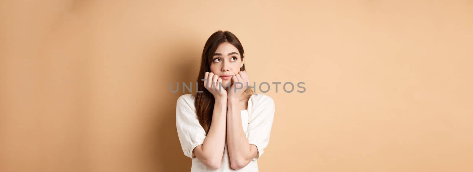 Dreamy cute woman looking pensive at upper left corner, thinking of something romantic, standing on beige background.