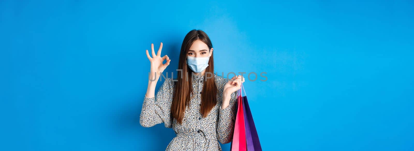 Covid-19, pandemic and lifestyle concept. Attractive woman wear medical mask on shopping, show okay and hold bags with purchases, blue background.
