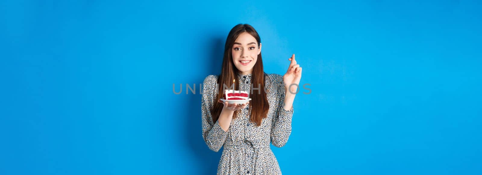 Hopeful birthday girl making wish on cake, cross fingers for good luck, standing in stylish dress against blue background by Benzoix