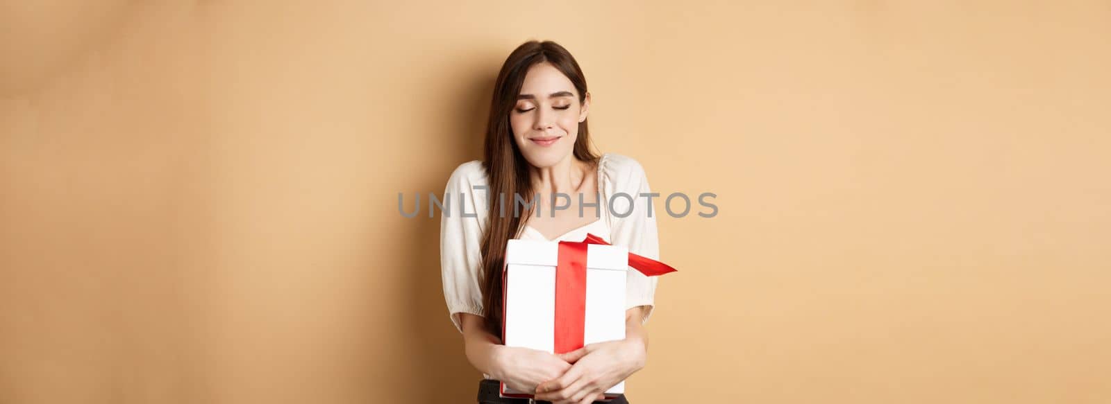 Happy valentines day. Dreamy woman hugging her surprise gift, close eyes and smile, standing on beige background by Benzoix