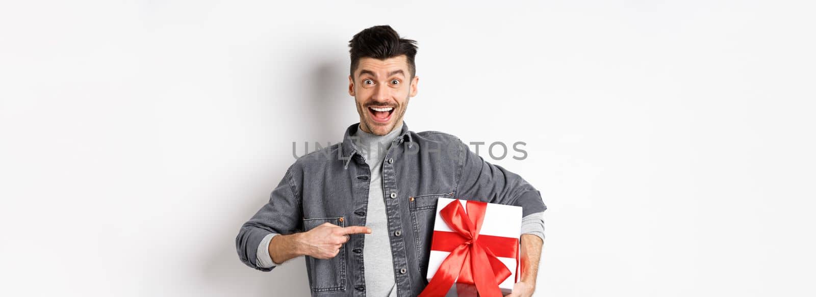 Happy Valentines day. Excited caucasian guy pointing at gift box from lover, smiling and looking amazed, buying presents on romantic date, white background by Benzoix