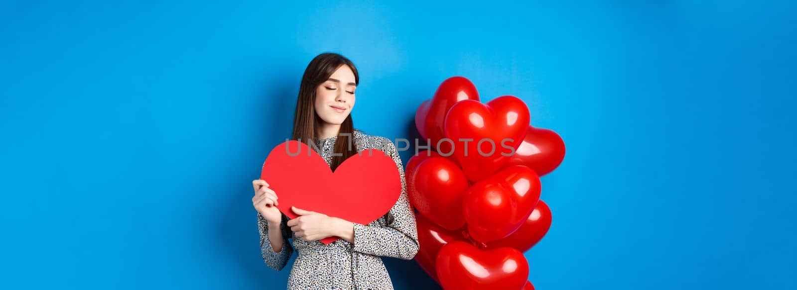 Valentines day. Romantic smiling woman hugging big red heart cutout and dreaming of love, standing in dress on blue background by Benzoix