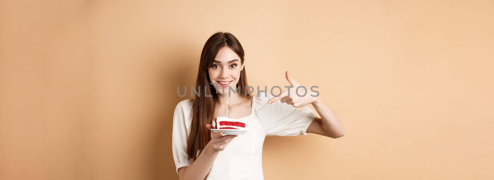 Cute girl pointing at birthday cake with candle and smiling excited, making bday wish, standing on beige background by Benzoix