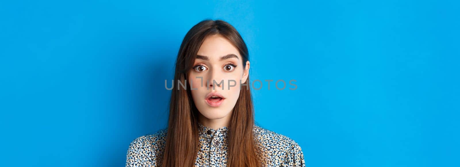 Close-up of excited pretty woman with long natural hair and makeup, drop jaw and gasping surprised, checking out awesome promo, hear amazing news, blue background.