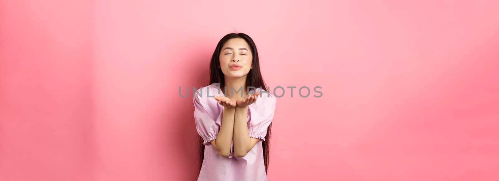 Cute and romantic asian girl blowing air kiss at camera, close eyes and smile tenderly, standing in dress against pink background by Benzoix