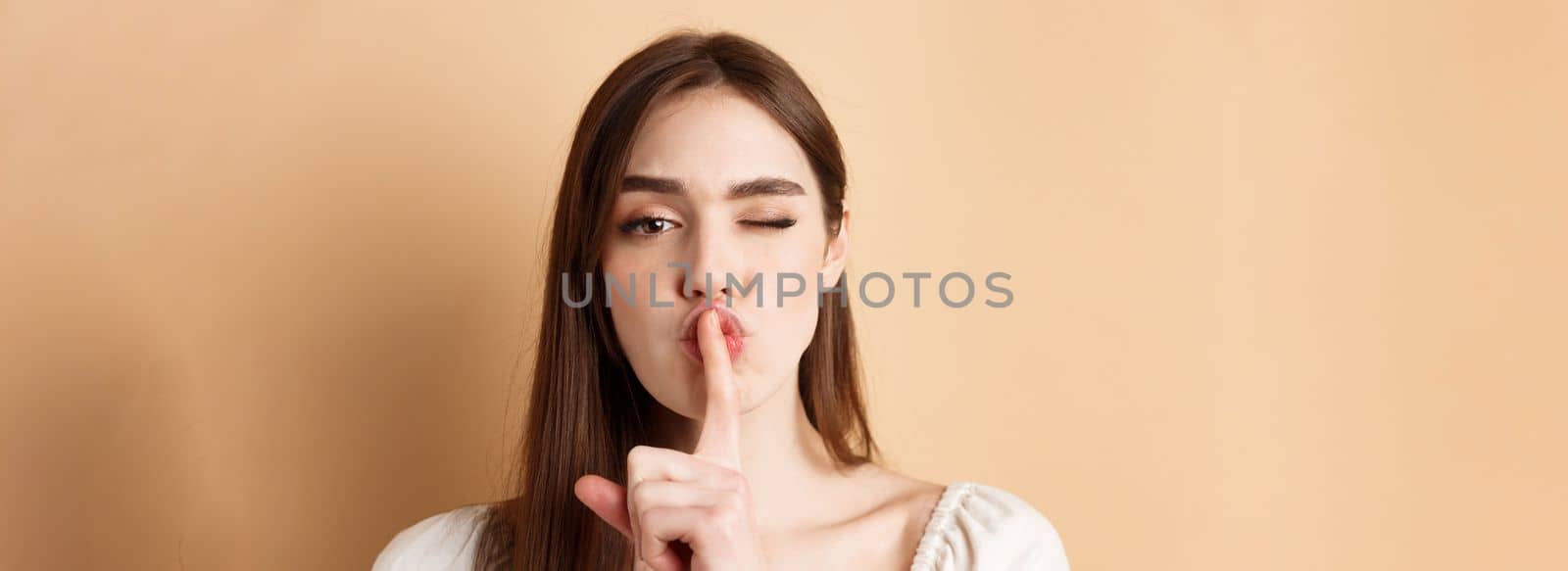 Close up of attractive woman shushing at camera, hiding secret, making hush sign and winking, show taboo sign, standing on beige background.