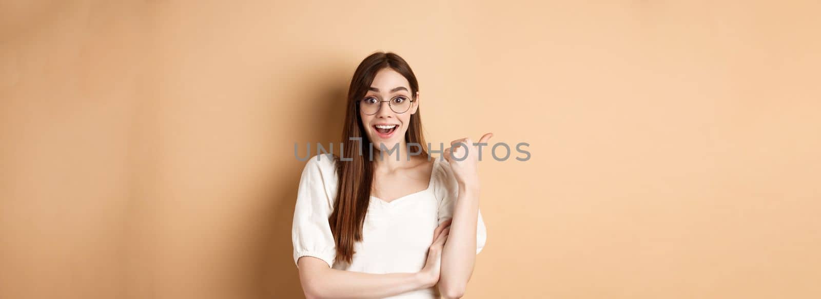 Excited girl in glasses pointing aside at logo, smiling amazed, recommending company, standing on beige background.
