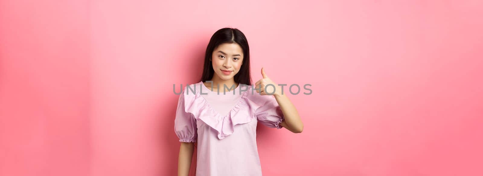 Smiling asian girl show thumb up in approval, praise good choice, like and recommend product, standing on pink background.