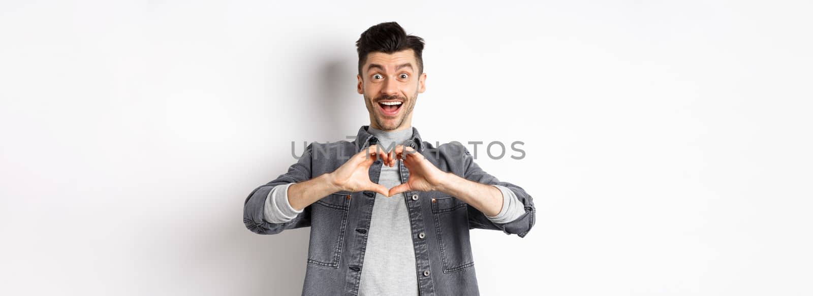 Romantic boyfriend showing heart gesture and looking with love at camera, celebrating White day after valentines, standing on white background. Romance concept.