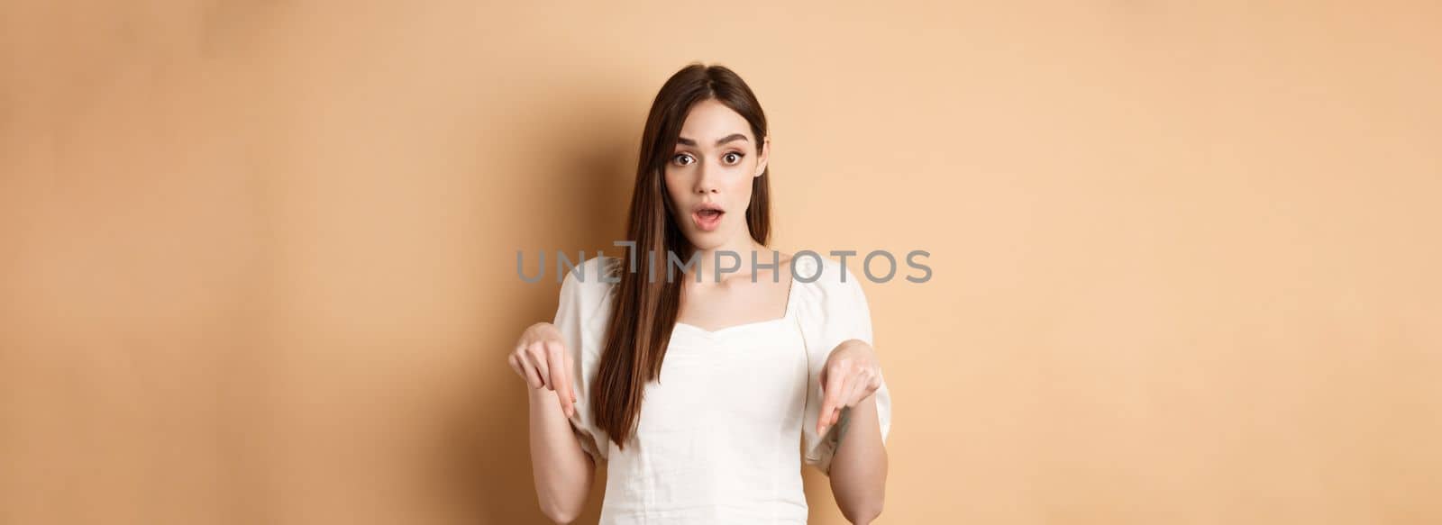 Wow look there. Amazed young woman in white dress pointing fingers down, standing in awe with dropped jaw and popped eyes against beige background.