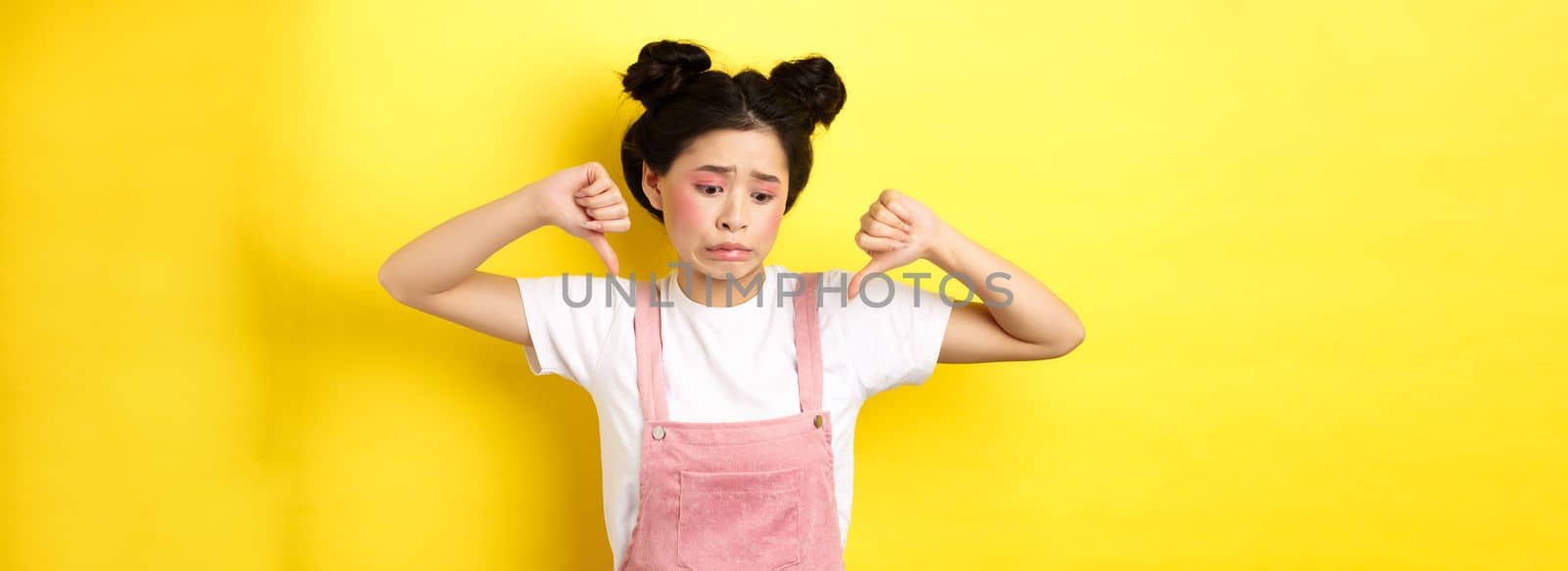 Asian beauty girl cringe from something disgusting, look away and showing thumbs-down to express dislike, negative emotions, standing in summer clothes on yellow background by Benzoix