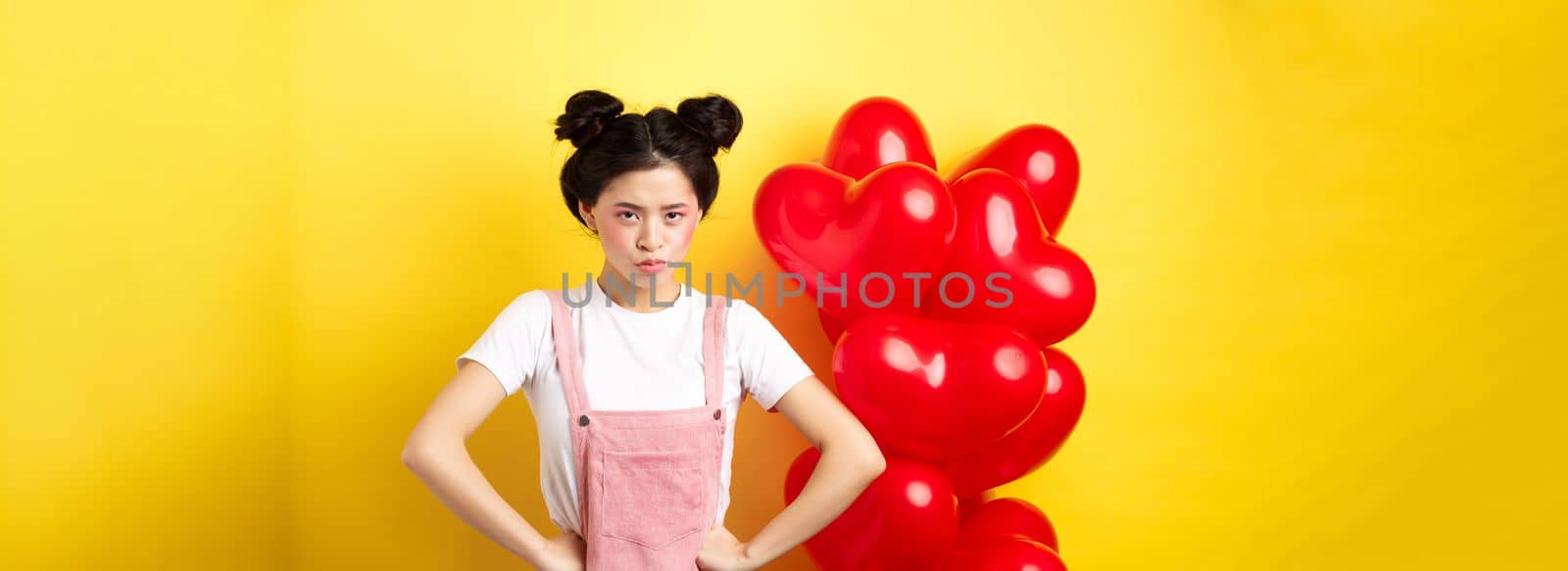 Valentines day and relationship concept. Disappointed asian girlfriend sulking, looking offended at camera, mad at boyfriend, standing near heart balloons, yellow background.