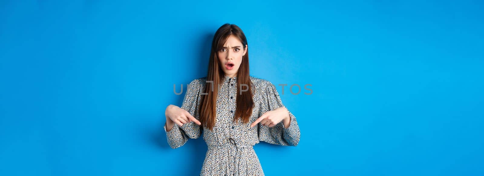 Shocked and offended young woman in dress frowning, gasping and pointing fingers down at insulting promo, standing on blue background.