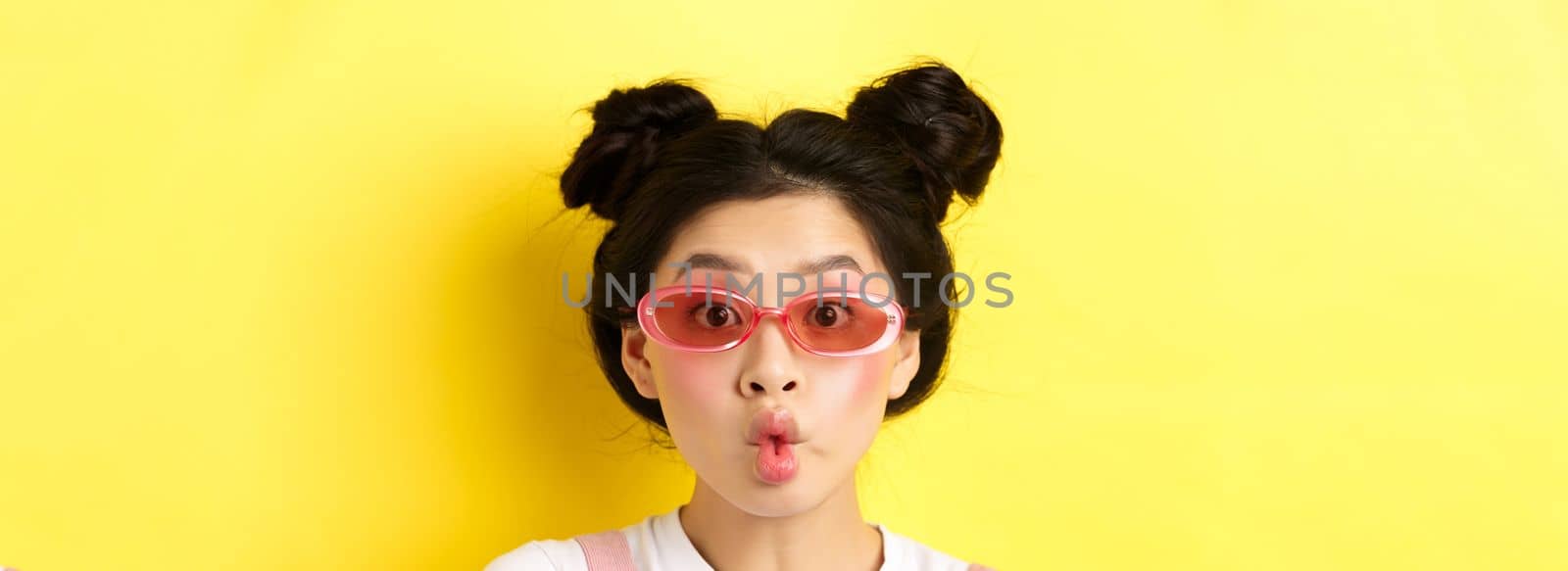Summer fashion concept. Silly glamour girl in sunglasses, pucker lips like fish and looking funny at camera, standing against yellow background.