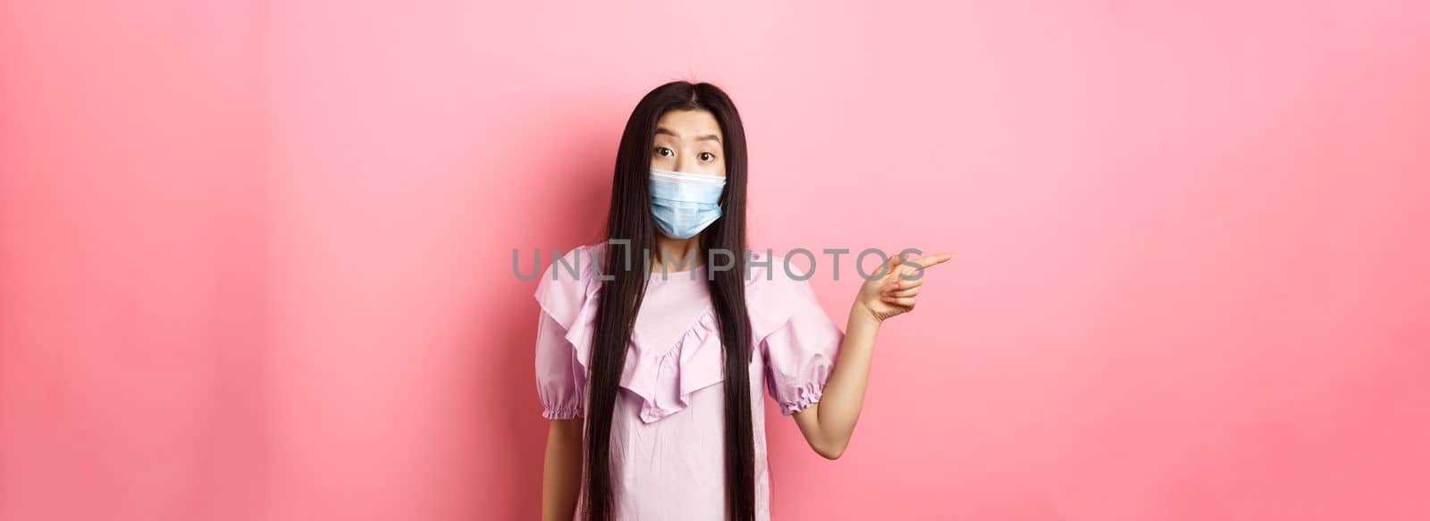 Covid-19, pandemic lifestyle concept. Curious asian girl in medical mask pointing left at logo, asking question about promotion, standing on pink background.
