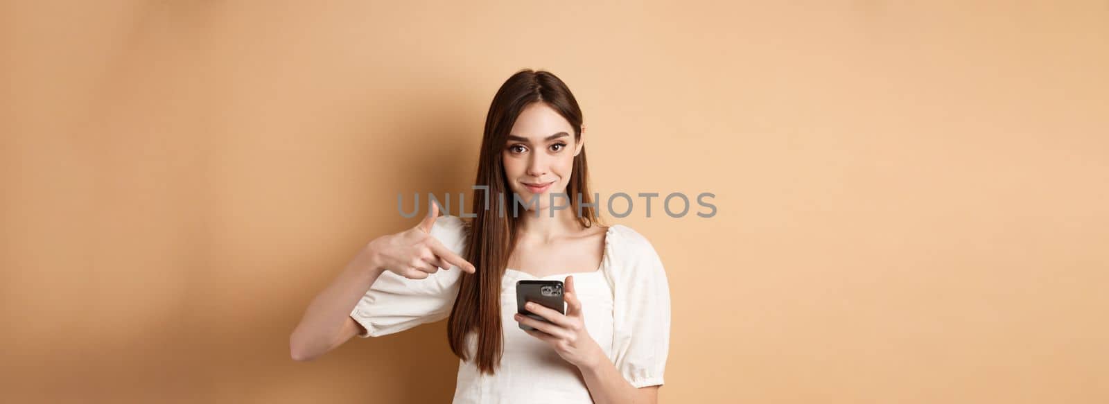 Attractive woman smiling and pointing at smartphone, showing online offer on phone, standing on beige background.