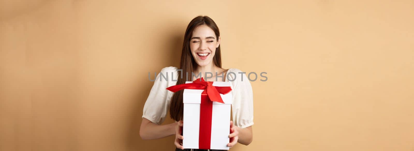 Happy smiling woman laughing, holding gift box from lover, enjoying romantic date on Valentines day, standing on beige background.