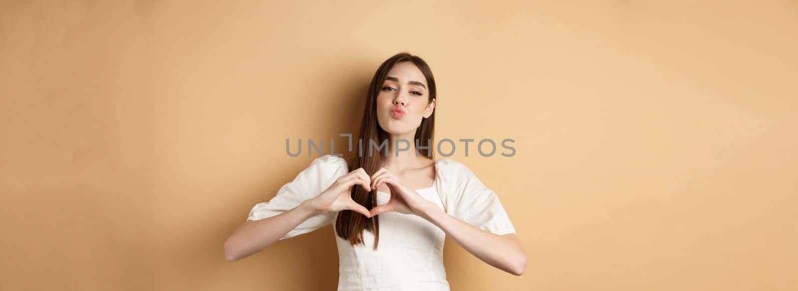 Valentines day. Lovely young womam in white dress, confess in love, pucker lips for kiss and showing heart gesture to lover, standing on beige background.