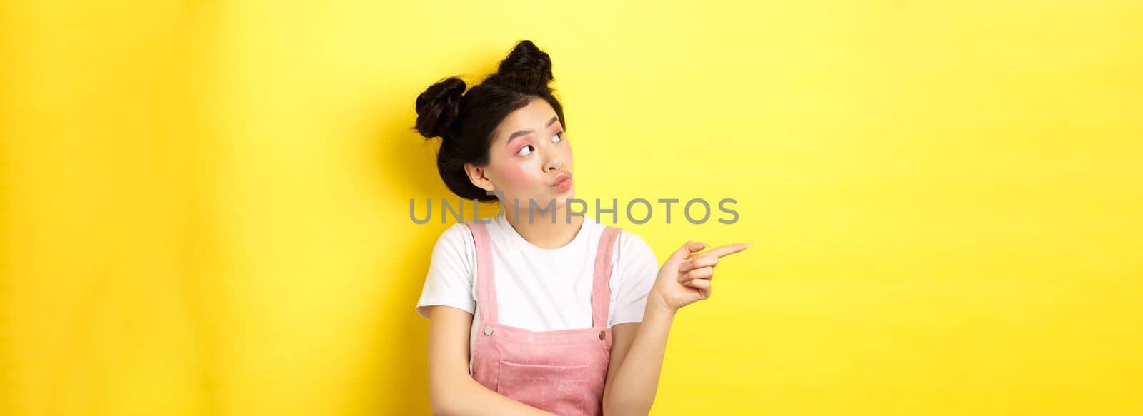 Pensive asian woman with beauty makeup, pointing and looking left with curious face, interested in advertisement, standing on yellow background.