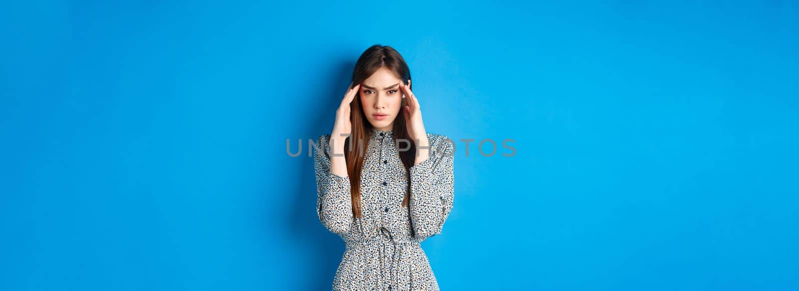 Woman trying to concentrate, touching head and squinting at camera, thinking or remember something, standing in dress on blue background.