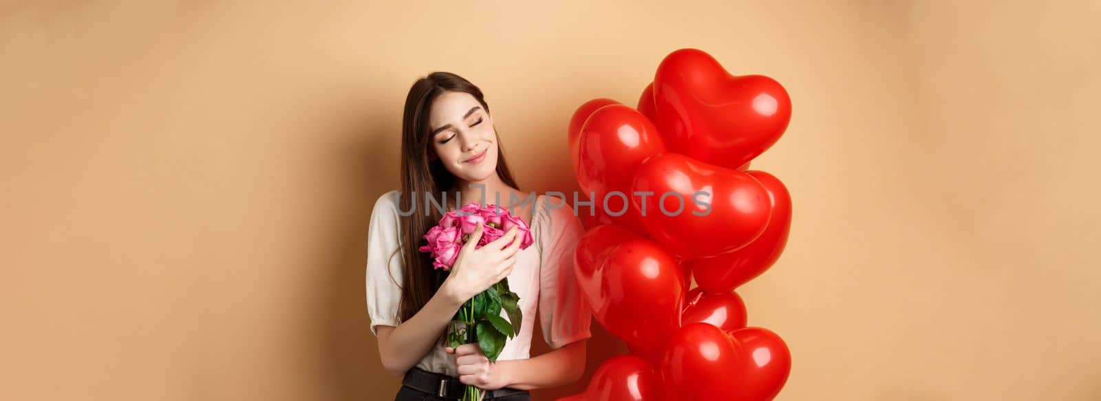 Sensual and romantic girl hugging bouquet of pink roses, smiling with eyes closed, thinking of lover, standing near Valentines day heart balloons, beige background by Benzoix