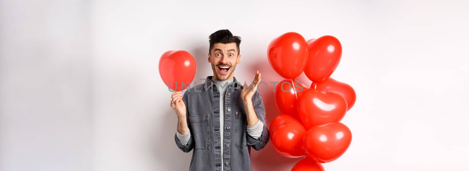 Valentines day concept. Cheerful young man celebrating love holiday, standing near red heart balloons and looking surprised, scream of joy, white background by Benzoix