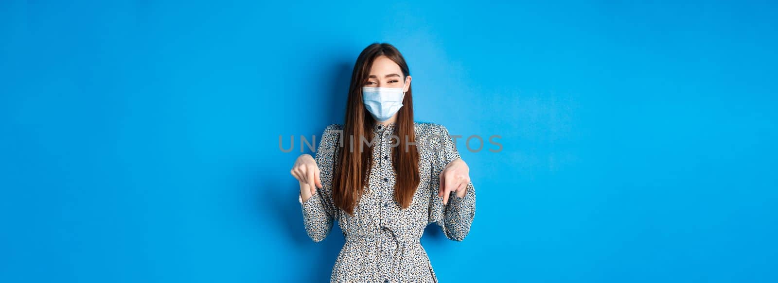 Covid-19, social distancing and healthcare concept. Happy beautiful person in medical mask pointing fingers down, laughing and smiling, showing advertisement, blue background by Benzoix