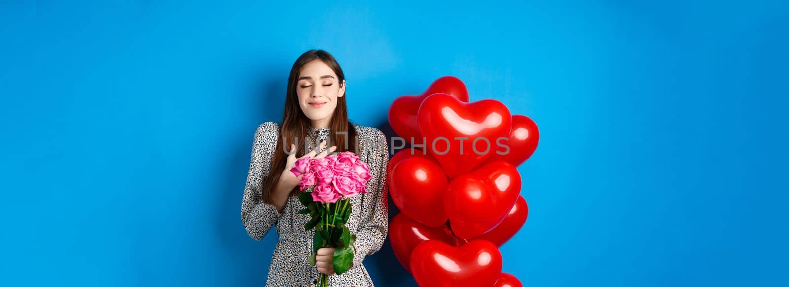 Valentines day. Romantic lovely woman in dress, close eyes and smiling, receiving flowers from lover, smelling bouquet of pink roses, blue background.