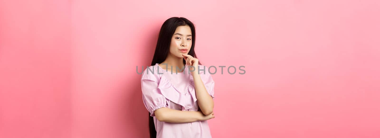 Hmm, interesting. Pensive and cunning asian girl having idea, plan to do something, looking at camera and touching chin with thoughtful face, standing on pink background.