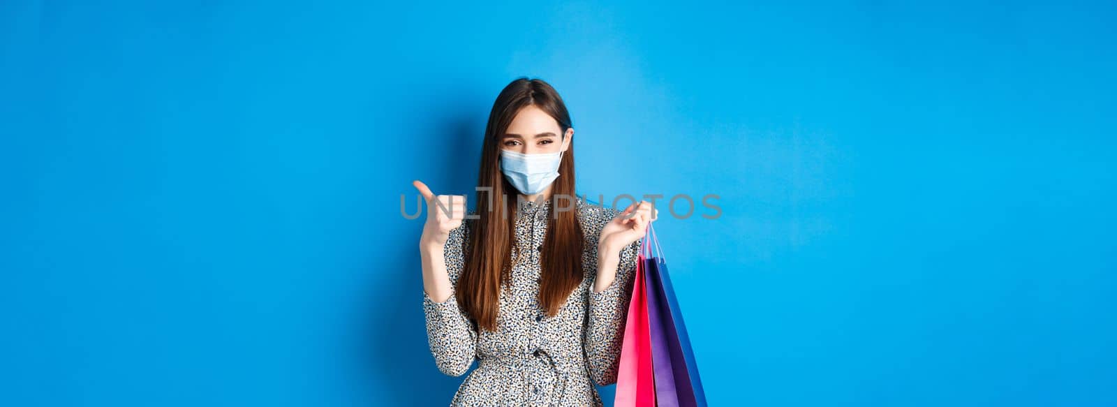 Covid-19, pandemic and lifestyle concept. Attractive woman wear medical mask on shopping, show thumb up and hold bags with purchases, blue background by Benzoix