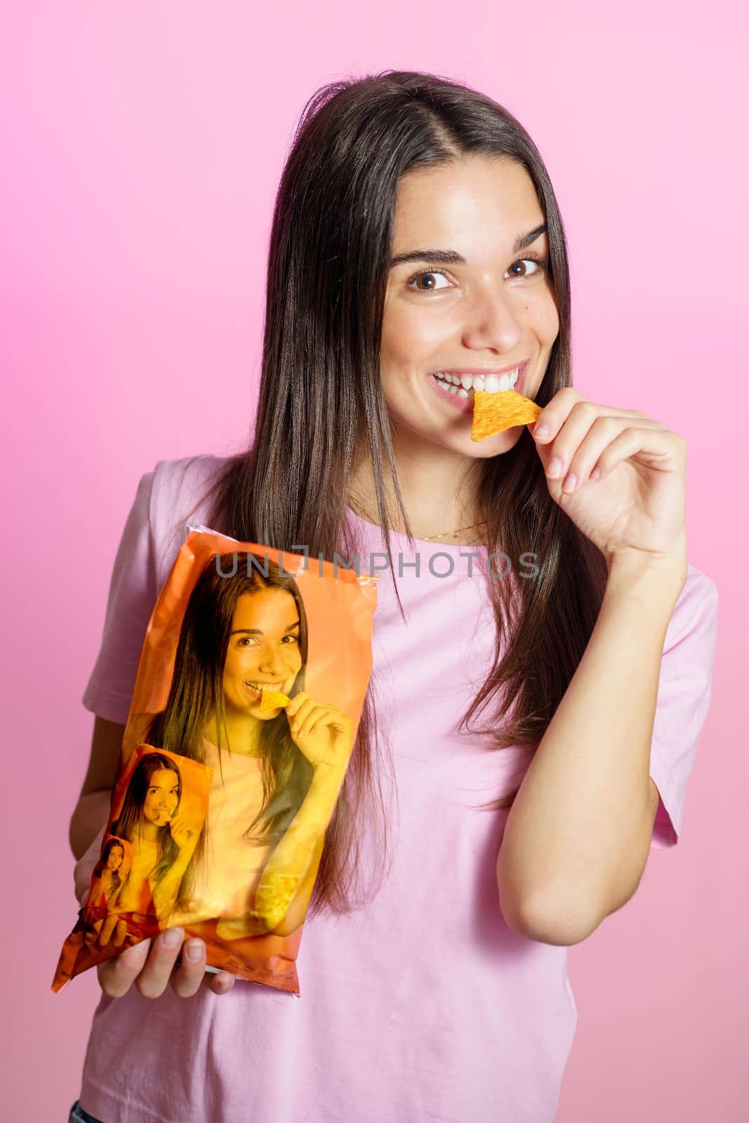 Young happy woman with long dark hair standing on pink background and biting delicious potato chip while holding package with own photos