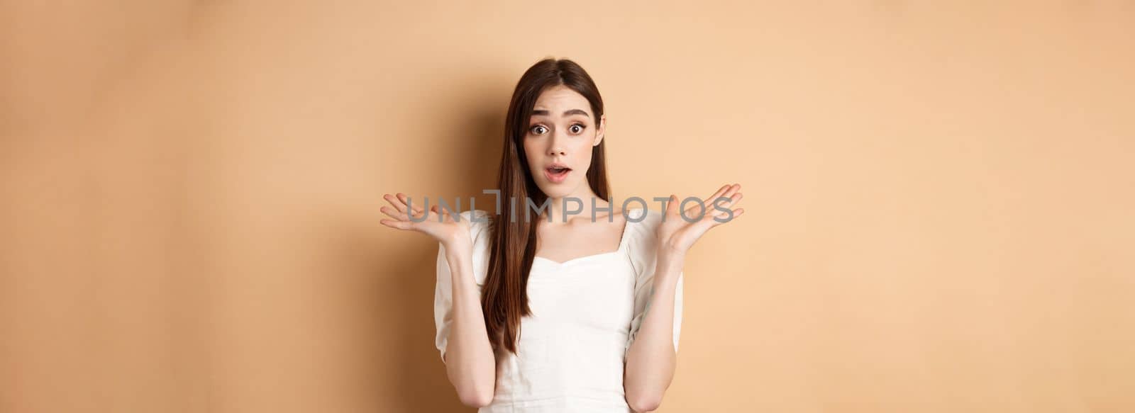 Surprised caucasian girl look with disbelief and amazement, spread hands sideways and gasping confused, cant understand what happening, know nothing, standing on beige background.