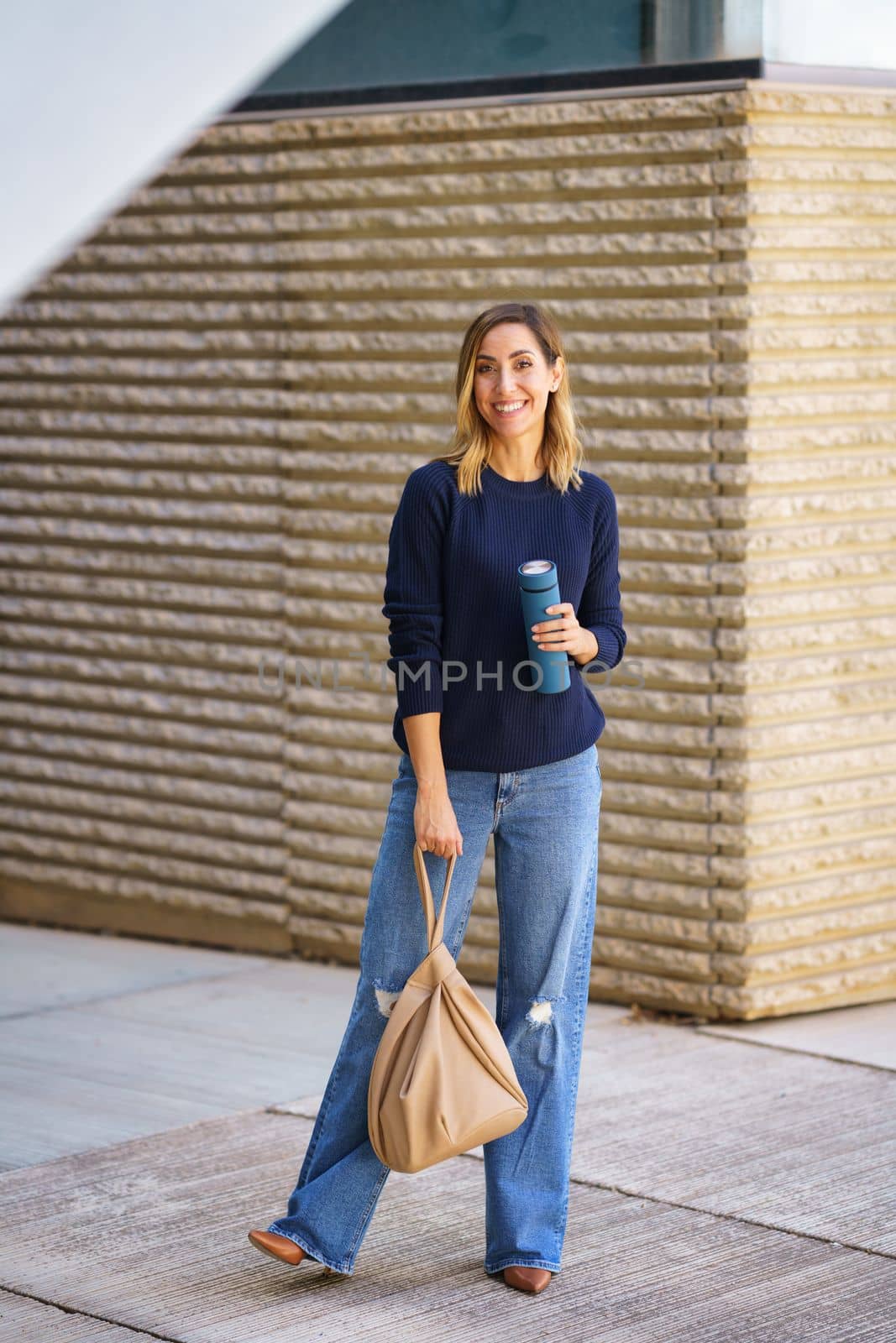 Full body cheerful woman in stylish outfit with bag and thermos standing on pavement and smiling near contemporary building on city street