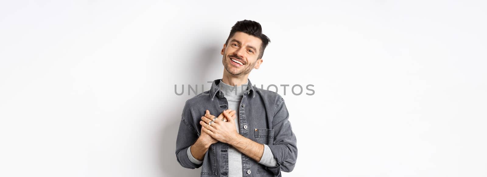 Romantic guy hold hands on heart and smiling, thanking you, looking pleased and grateful, standing on white background by Benzoix