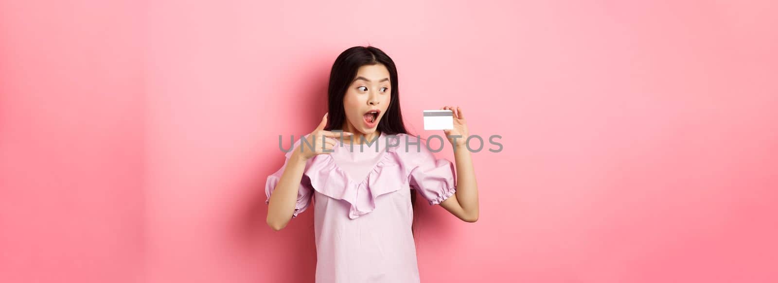 Excited asian woman pointing at plastic credit card, showing awesome advertisement, standing on pink background.