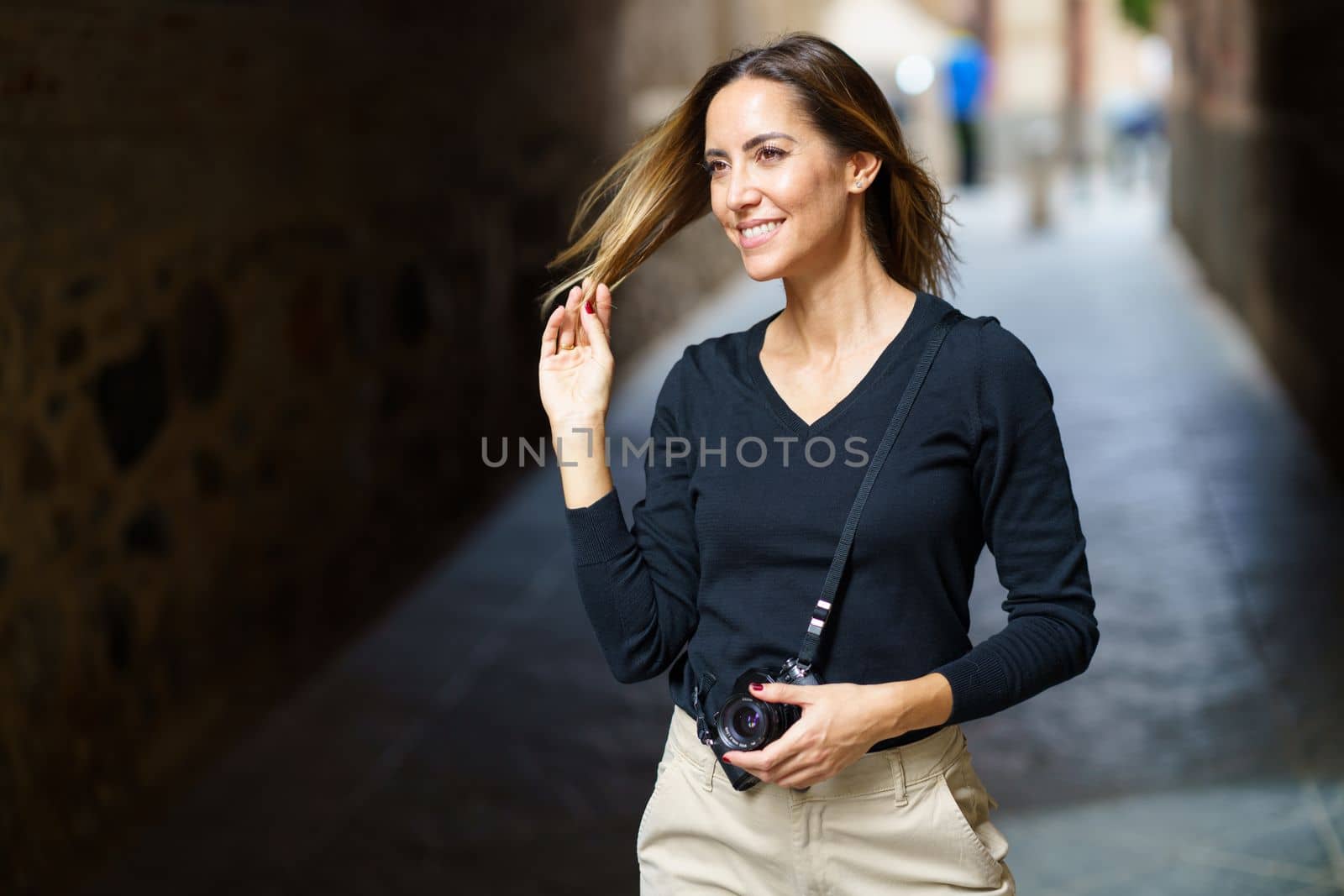 Content young female tourist in casual wear with retro photo camera, touching long brown hair and smiling while standing in narrow paved street in old town