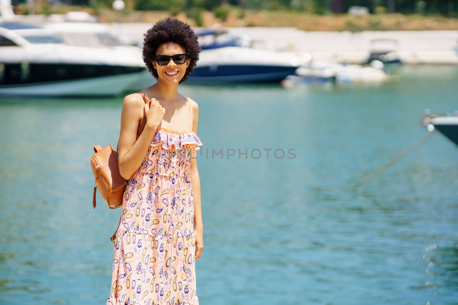 Happy black woman in dress and sunglasses with bag, on shoulder smiling and looking at camera on blurred background on sea and boats on summer day in harbor