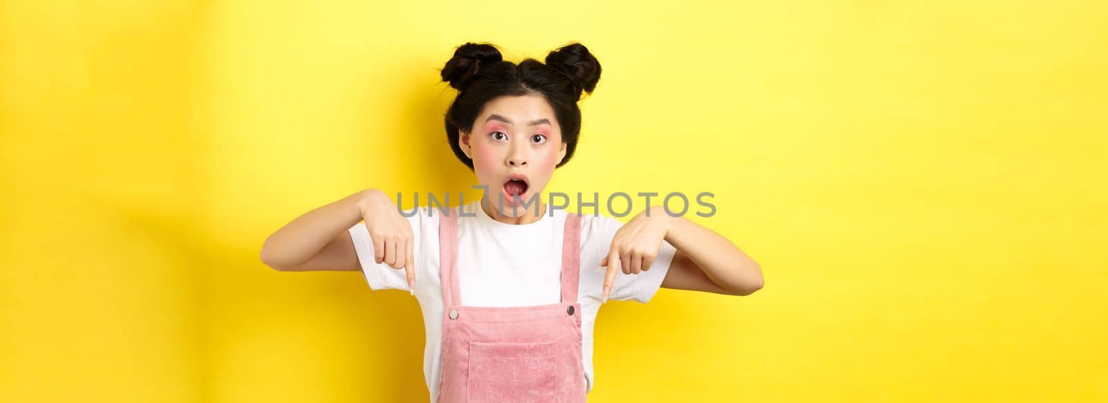 Surprised asian woman with glamour makeup and hairstyle, drop jaw and pointing down amazed, standing against yellow background.