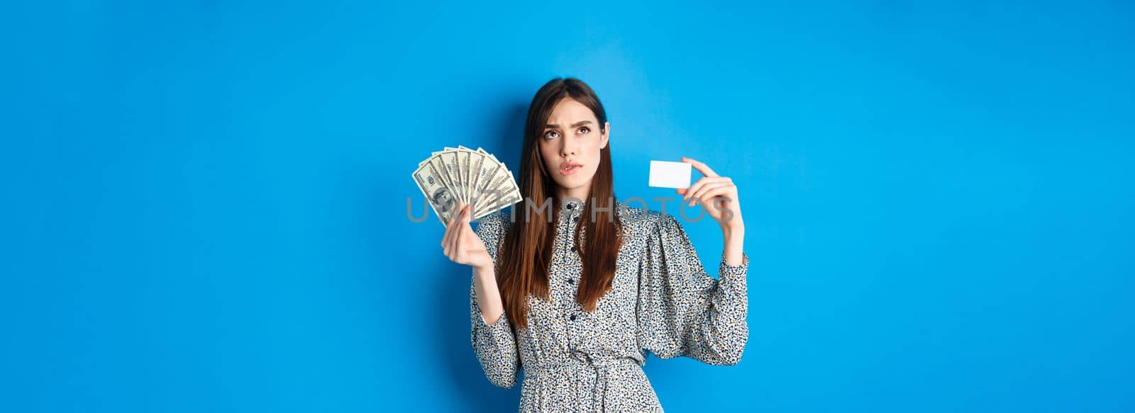 Shopping. Thoughtful girl looking up and biting lip, counting in mind or thinking, holding plastic credit card with dollar bills, standing on blue background.